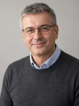 Portret al lui Radu Iordache, cu ochelari și pulover gri peste cămașă albastră, pe fundal gri