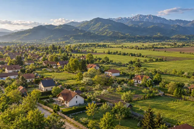 Vedere din dronă peste loturi verzi și case noi la marginea comunei, cu munți în spate